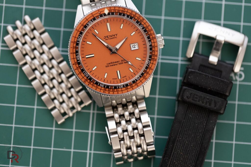 Reloj Jenny Caribbean 300 con esfera naranja, brazalete de acero beads of rice y correa de caucho negra sobre base de corte verde para guía de medidas de muñeca y correas.