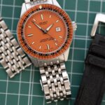 Reloj Jenny Caribbean 300 con esfera naranja, brazalete de acero beads of rice y correa de caucho negra sobre base de corte verde para guía de medidas de muñeca y correas.