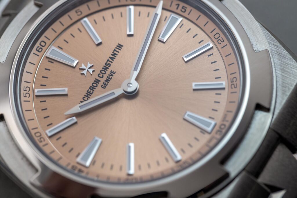 Macrofotografía del dial cepillado en tono salmón de un Vacheron Constantin Overseas, mostrando el control de luz y reflejos en cristal de zafiro.